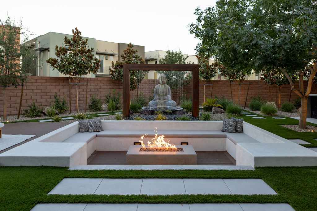 A modern backyard featuring a fire pit, seating area, Buddha statue, and water and fire features for a serene atmosphere.