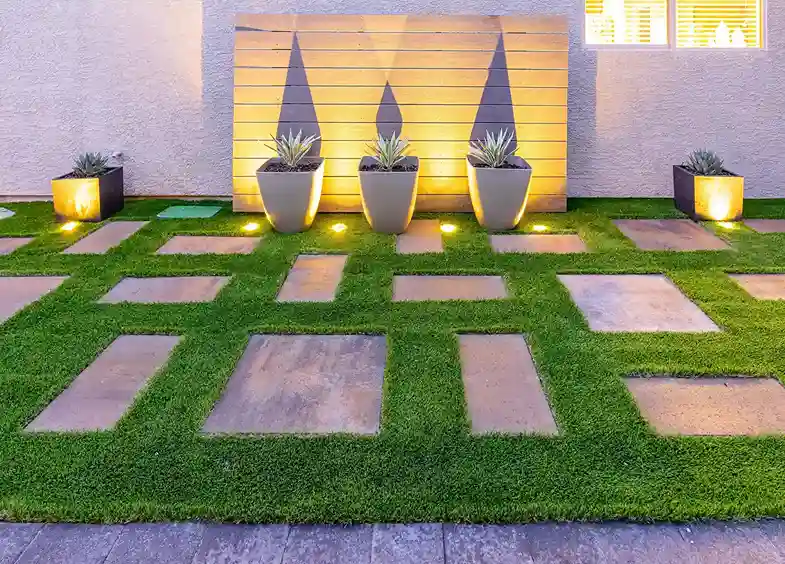 Modern garden with geometric stone path on green grass, three potted plants against a wooden panel, and soft warm landscape lighting.