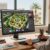A modern desktop setup showcasing advanced landscape design software. In the foreground, a sleek computer monitor displays a vibrant, intricate 3D landscape plan featuring lush gardens, elegant pathways, and various plant designs. A professional landscape designer, dressed in smart casual attire, sits focused at the desk, interacting with the software using a digital pen. The middle layer includes a neatly organized workspace with design tools, color swatches, and landscape books. In the background, large windows reveal a sunny Las Vegas skyline, with cactus and palm trees lining the view. The scene is illuminated by warm, natural light, creating an inviting atmosphere that embodies creativity and professionalism. Capture an angle that emphasizes both the designer's engagement and the software's dynamic features.